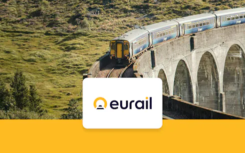 Train crossing a stone viaduct in a scenic European landscape.