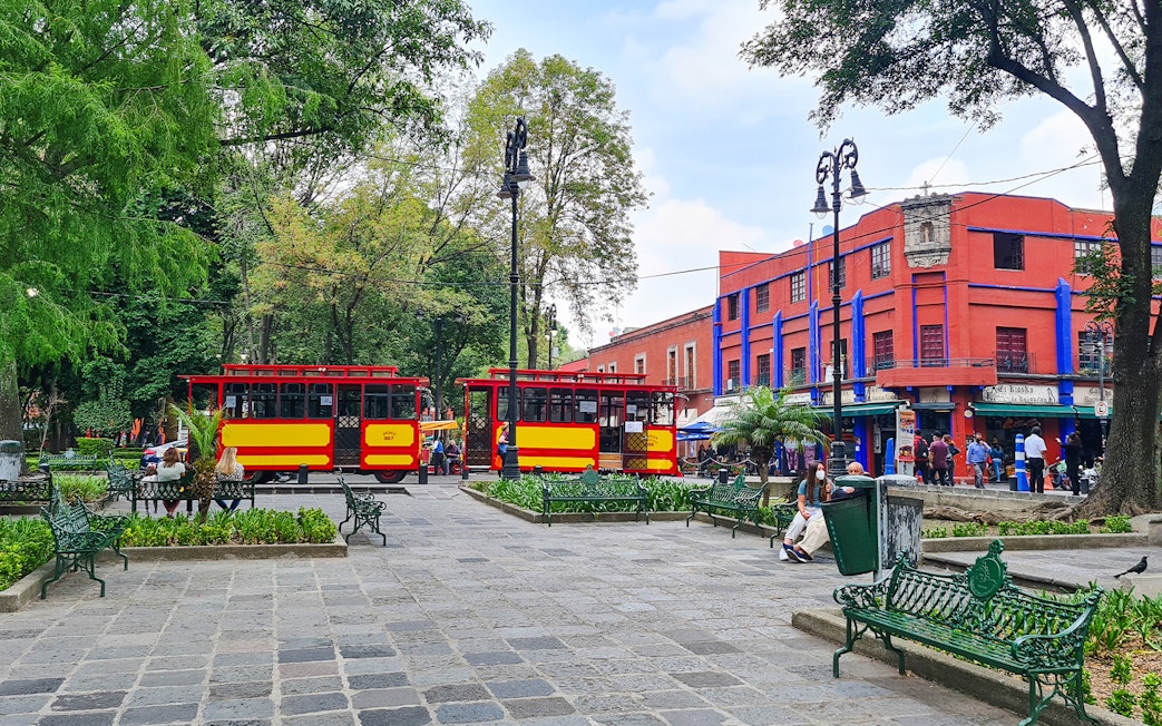 Red and yellow trams in Coyoacán square, Mexico City, with vibrant buildings and trees.