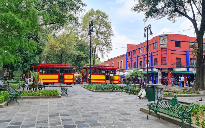 Red and yellow trams in Coyoacán square, Mexico City, with vibrant buildings and trees.