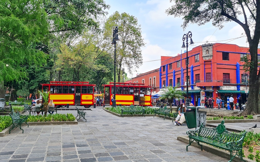 Red and yellow trams in Coyoacán square, Mexico City, with vibrant buildings and trees.