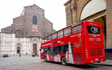 Red City tour bus in Bologna near historic church, part of hop-on hop-off tour with food tasting vouchers.