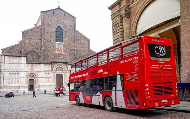 Red City tour bus in Bologna near historic church, part of hop-on hop-off tour with food tasting vouchers.