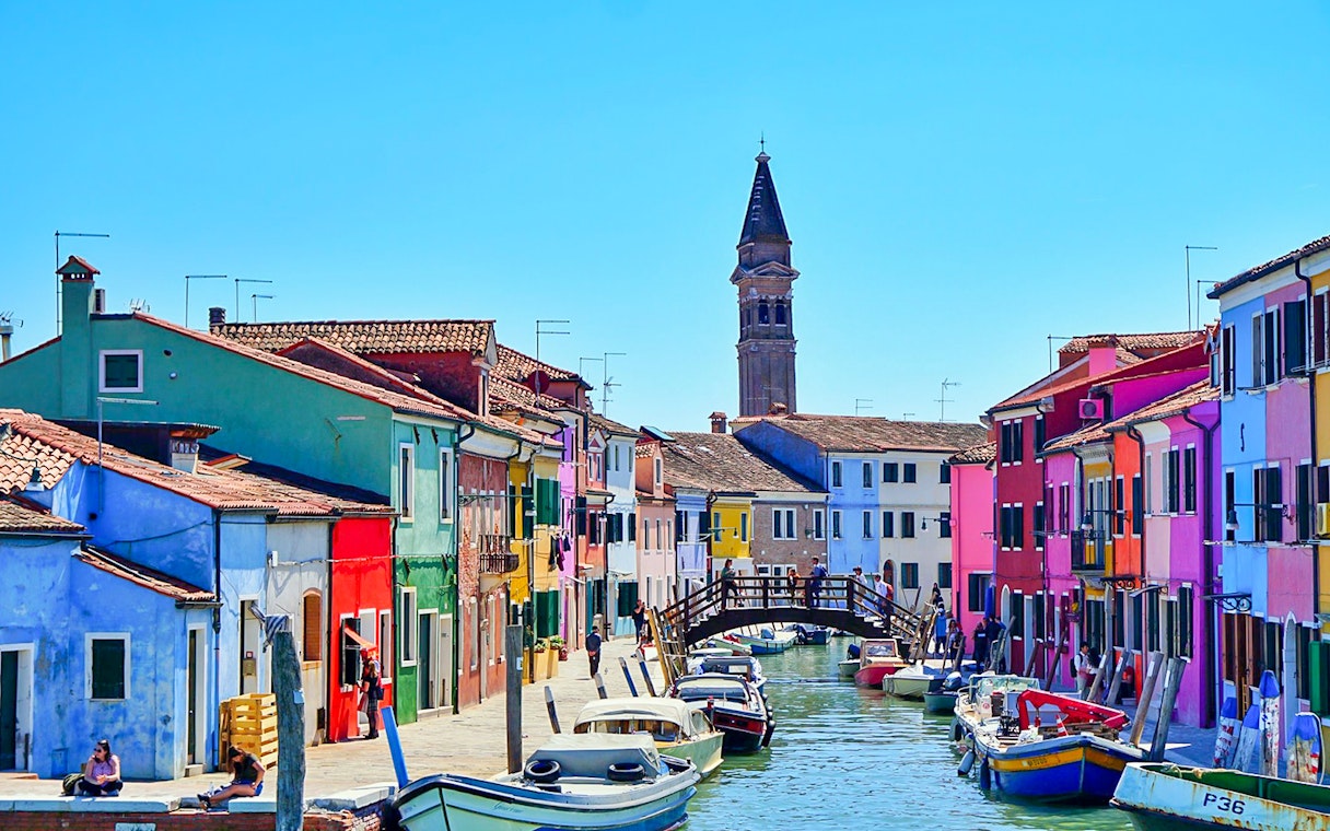Colorful houses along a canal in Burano, Venice, with boats and a wooden bridge.