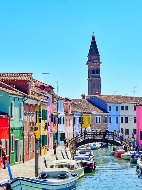 Colorful houses along a canal in Burano, Venice, with boats and a wooden bridge.