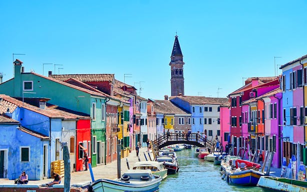 Colorful houses along a canal in Burano, Venice, with boats and a wooden bridge.