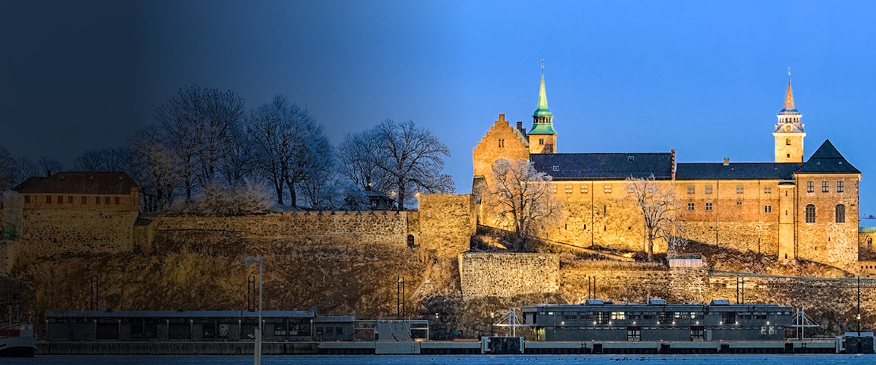 Akershus Fortress in Oslo, Norway, illuminated at dusk with bare trees in the background.