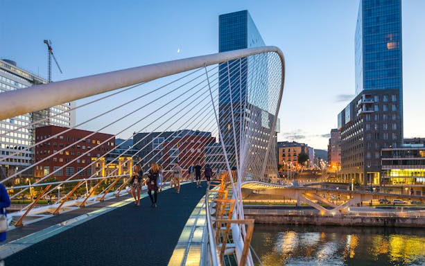 Pedestrians on Zubizuri Bridge in Bilbao at dusk, with cityscape and river views.