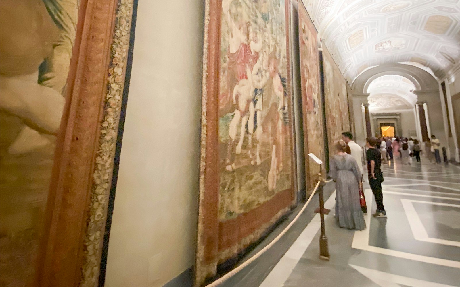 Tapestry gallery in the Vatican Museums with visitors observing artwork.