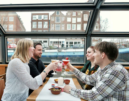 Friends enjoying cocktails on an Amsterdam canal cruise with city view.