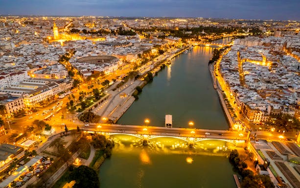 Aerial view of Seville's Triana district at night, featuring the illuminated Triana Bridge over the Guadalquivir River.
