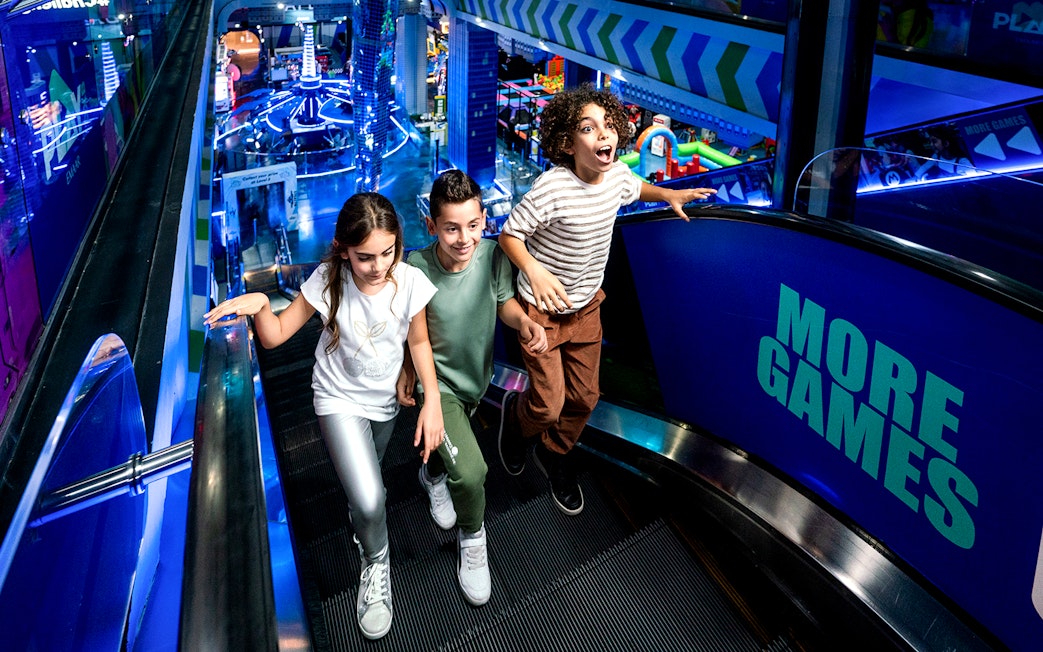 Children on escalator at Play DXB, Dubai, with arcade games in background.