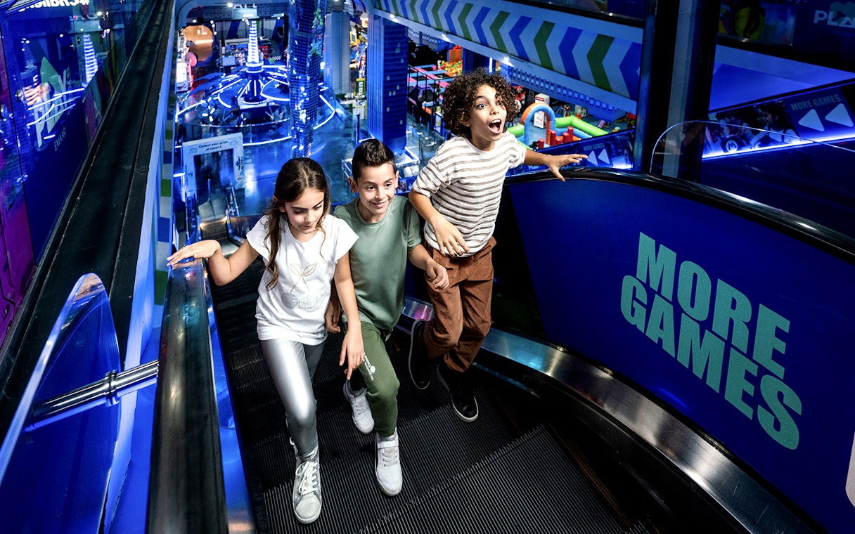 Children on escalator at Play DXB, Dubai, with arcade games in background.
