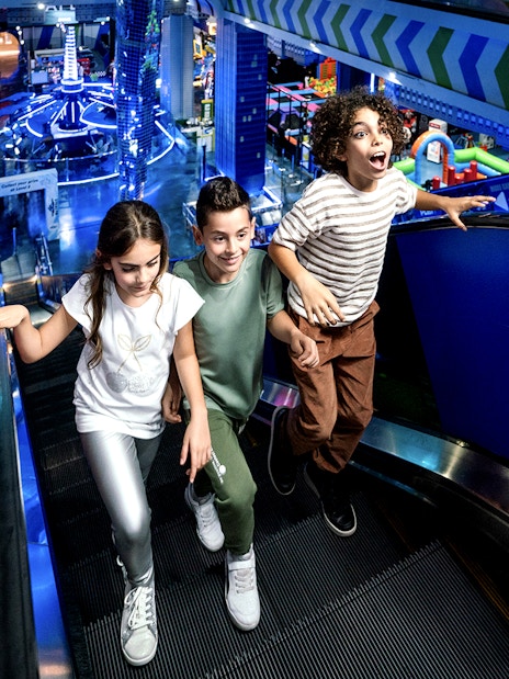 Children on escalator at Play DXB, Dubai, with arcade games in background.
