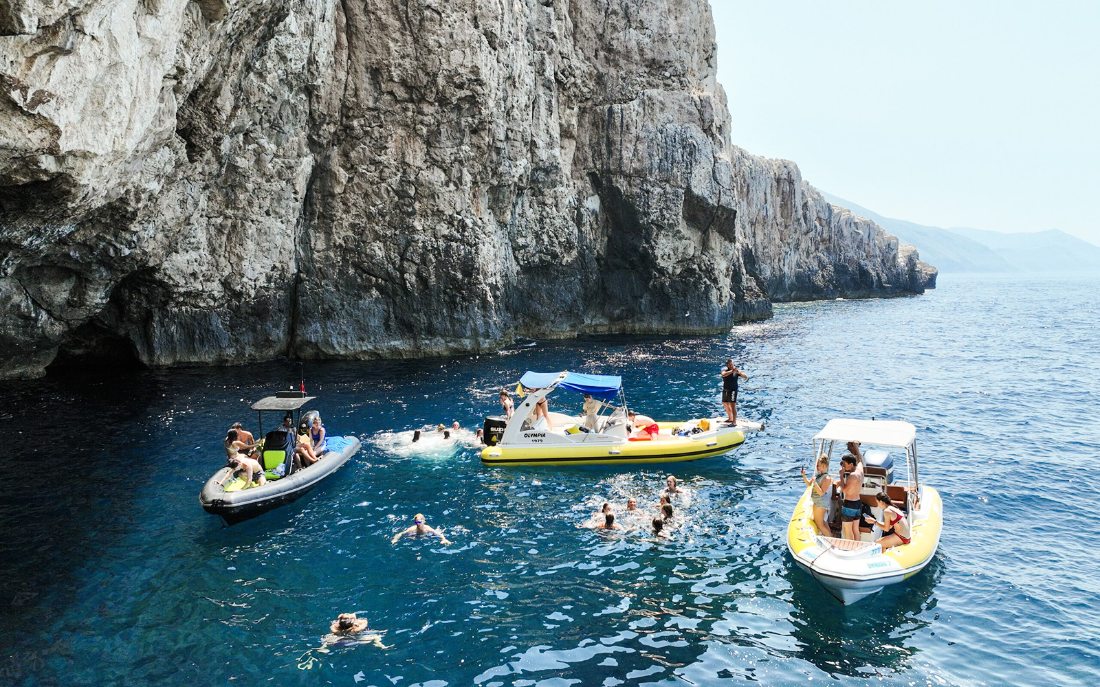 Small boats and swimmers near rocky cliffs at Dafina Bay and Haxhi Ali Cave.