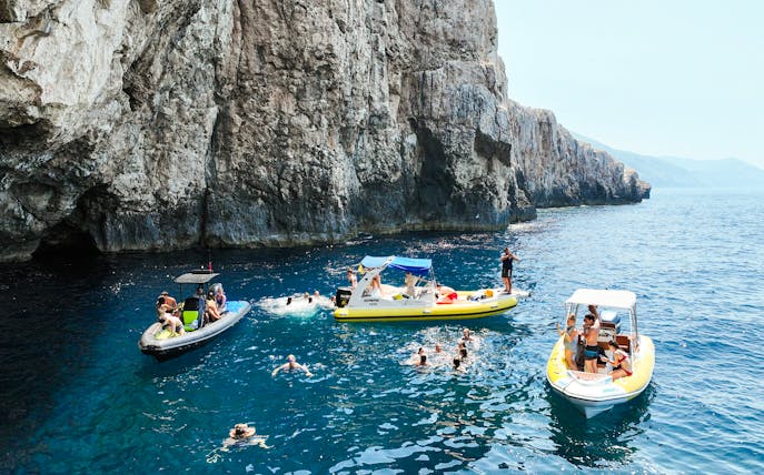 Small boats and swimmers near rocky cliffs at Dafina Bay and Haxhi Ali Cave.