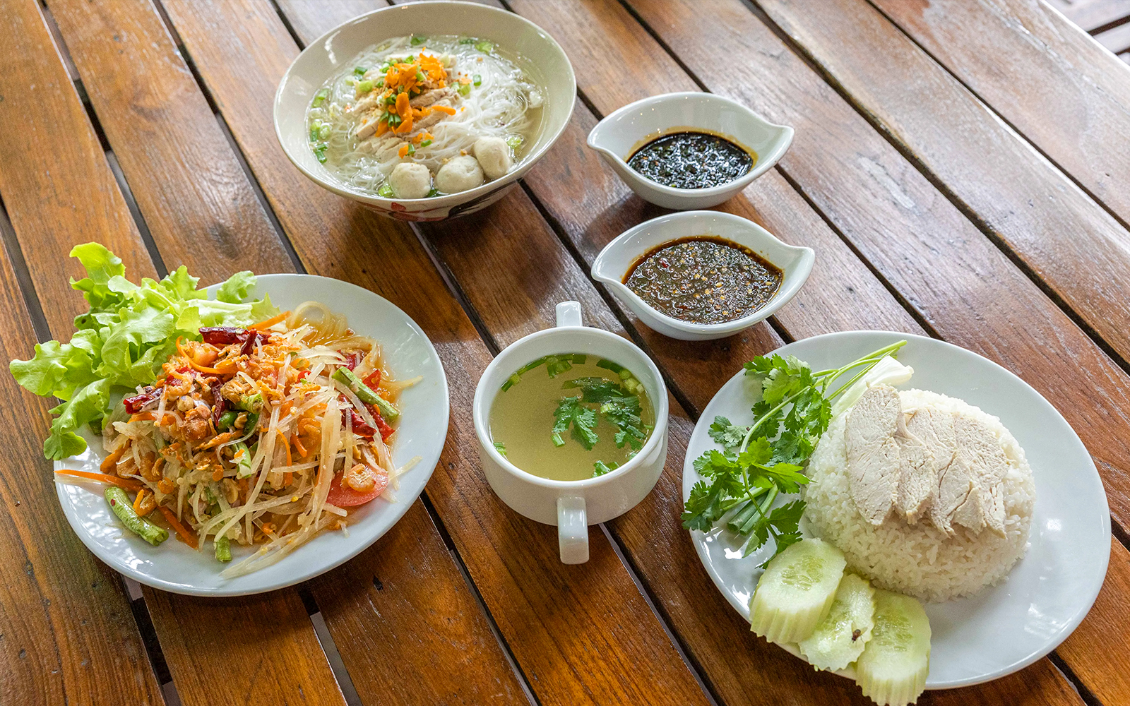 Traditional Thai meal with papaya salad, chicken rice, noodle soup, and sauces on a wooden table.