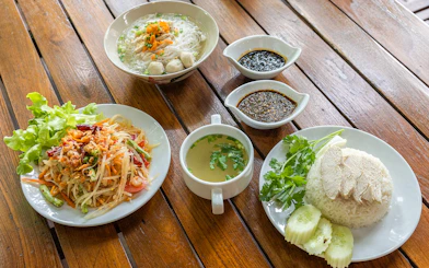Traditional Thai meal with papaya salad, chicken rice, noodle soup, and sauces on a wooden table.