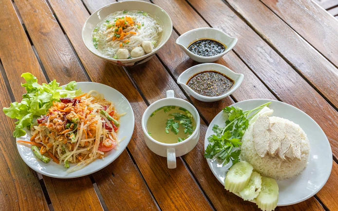 Traditional Thai meal with papaya salad, chicken rice, noodle soup, and sauces on a wooden table.
