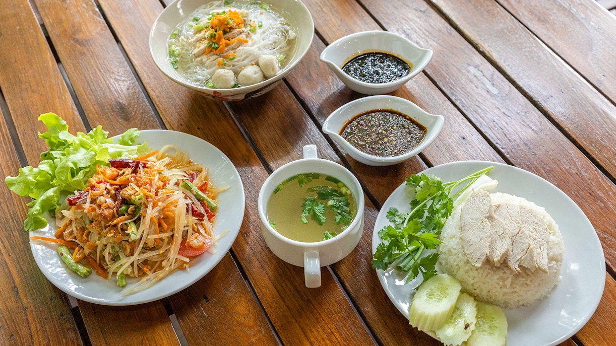Traditional Thai meal with papaya salad, chicken rice, noodle soup, and sauces on a wooden table.