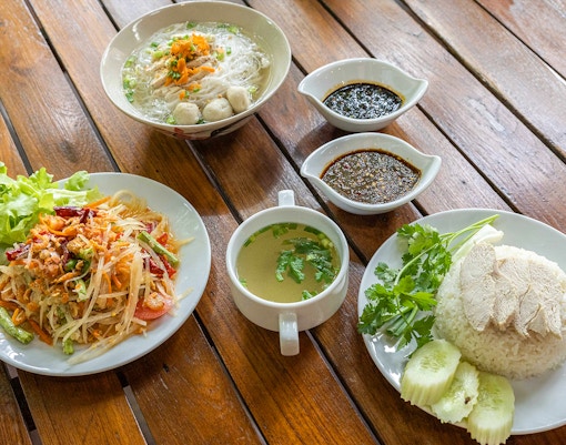 Traditional Thai meal with papaya salad, chicken rice, noodle soup, and sauces on a wooden table.