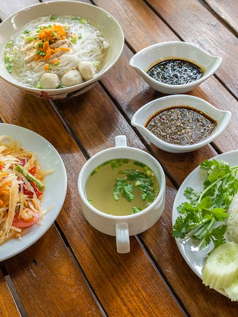 Traditional Thai meal with papaya salad, chicken rice, noodle soup, and sauces on a wooden table.