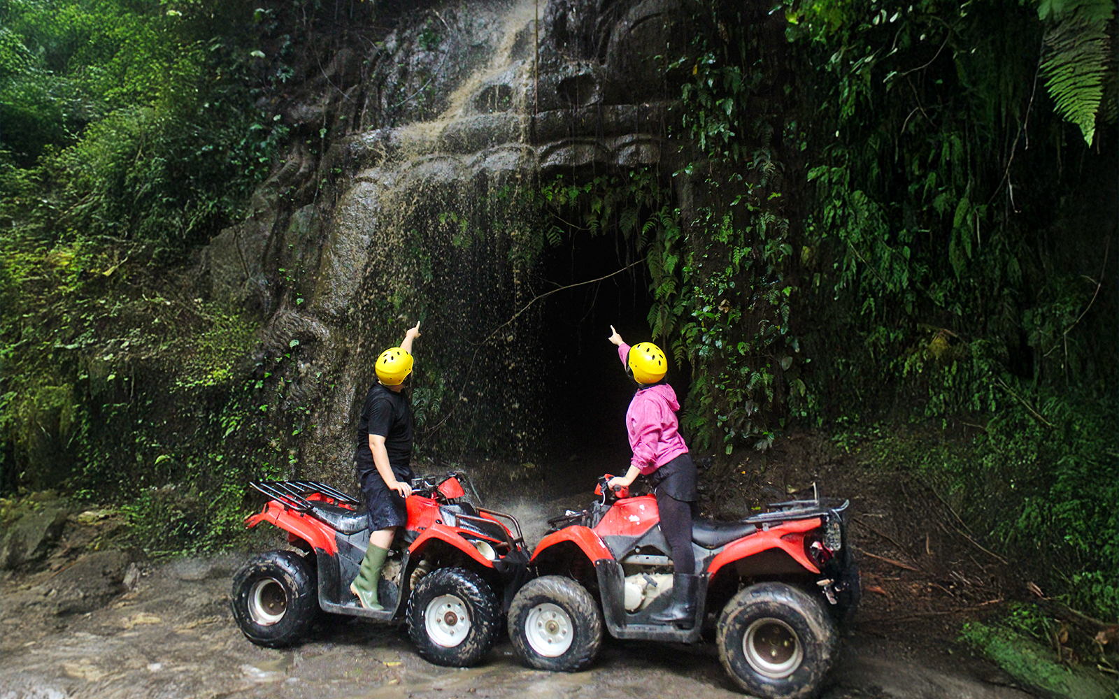 Two people on ATVs pointing at a cave entrance in a lush Bali jungle.