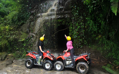 Two people on ATVs pointing at a cave entrance in a lush Bali jungle.