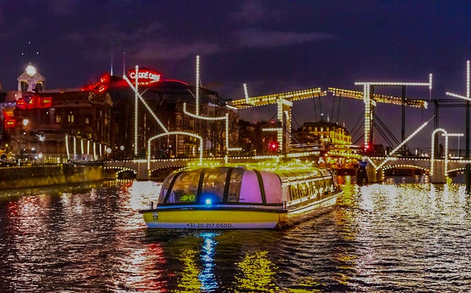 Canal boat cruising under illuminated bridges during Amsterdam Lights Festival.