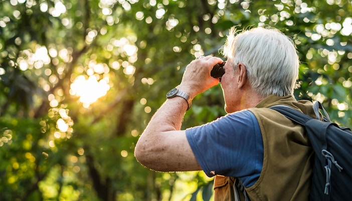 Elderly man using binoculars for bird watching in a lush green park.