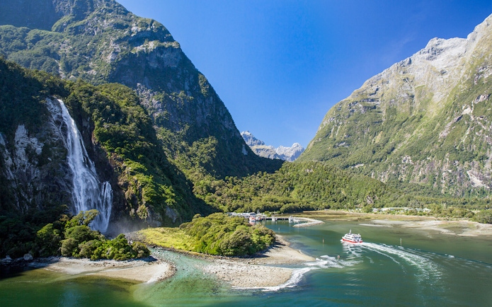 Cruise boat navigating Milford Sound with waterfall and lush mountains in New Zealand.