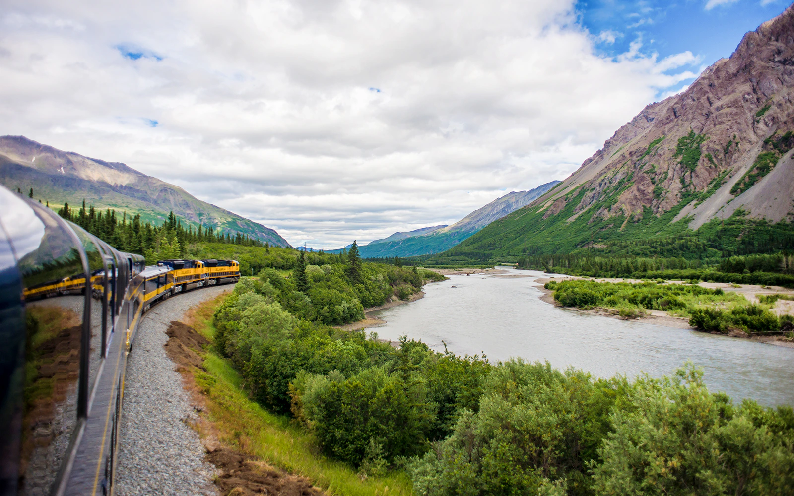 Alaska Railroad train winding through forest with snow-capped mountains in the background.