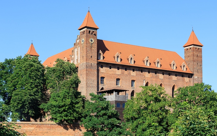 Medieval Teutonic brick castle with towers and trees in foreground.
