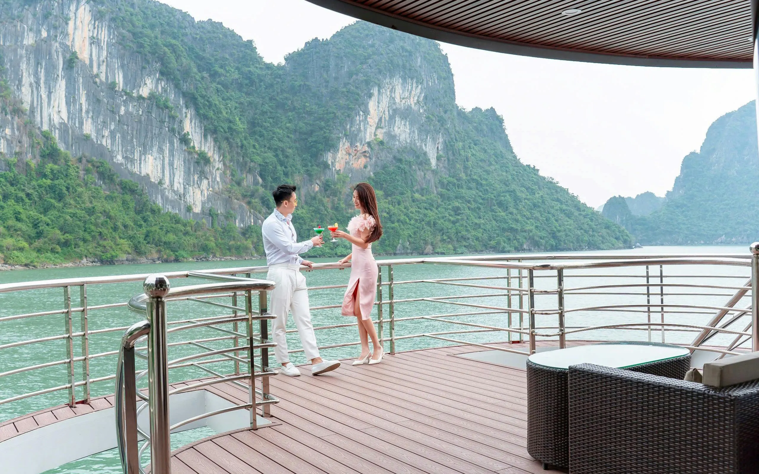 Couple enjoying drinks on Ha Long Bay luxury cruise deck with limestone cliffs in view.