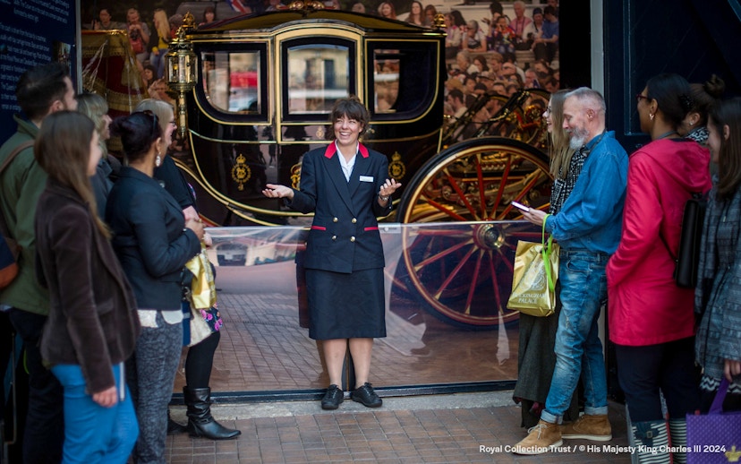 Warden guiding family visitors at The Royal Mews, London, with a historic carriage in the background.