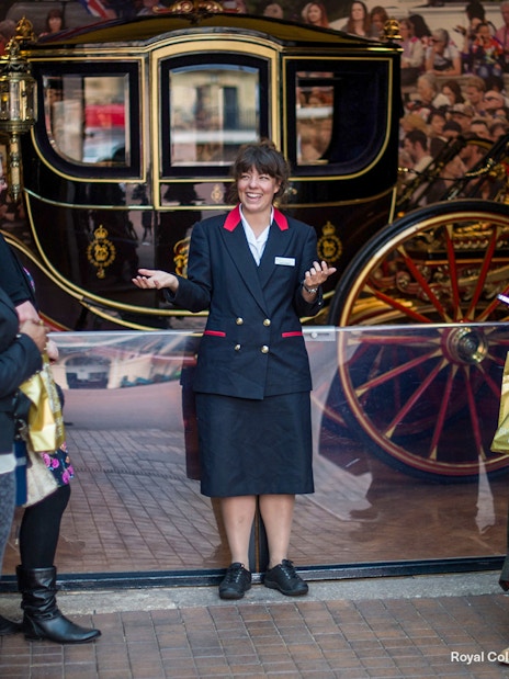 Warden guiding family visitors at The Royal Mews, London, with a historic carriage in the background.