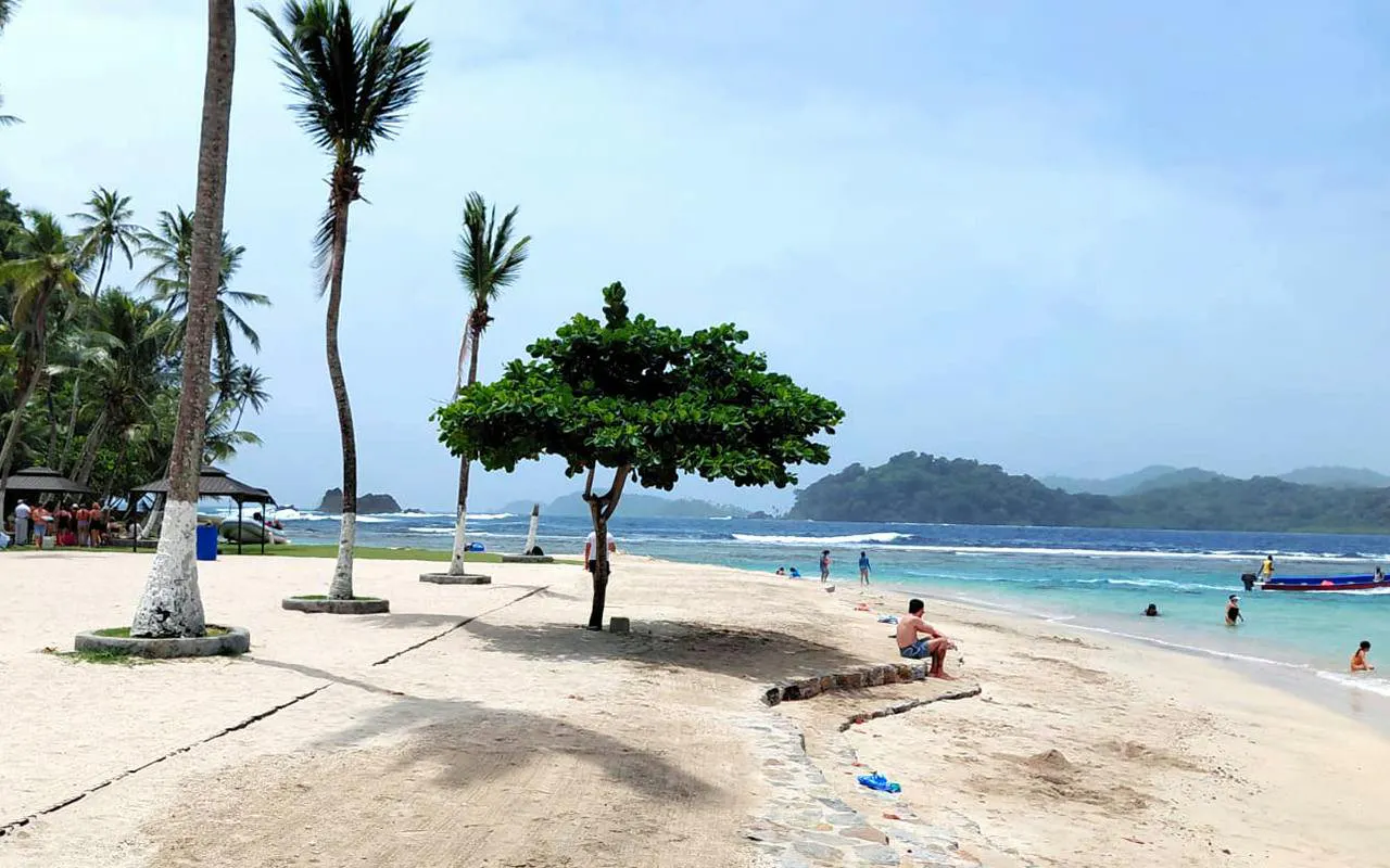 Beach scene at Isla Mamey with people relaxing and swimming, palm trees, and distant hills in Panama.