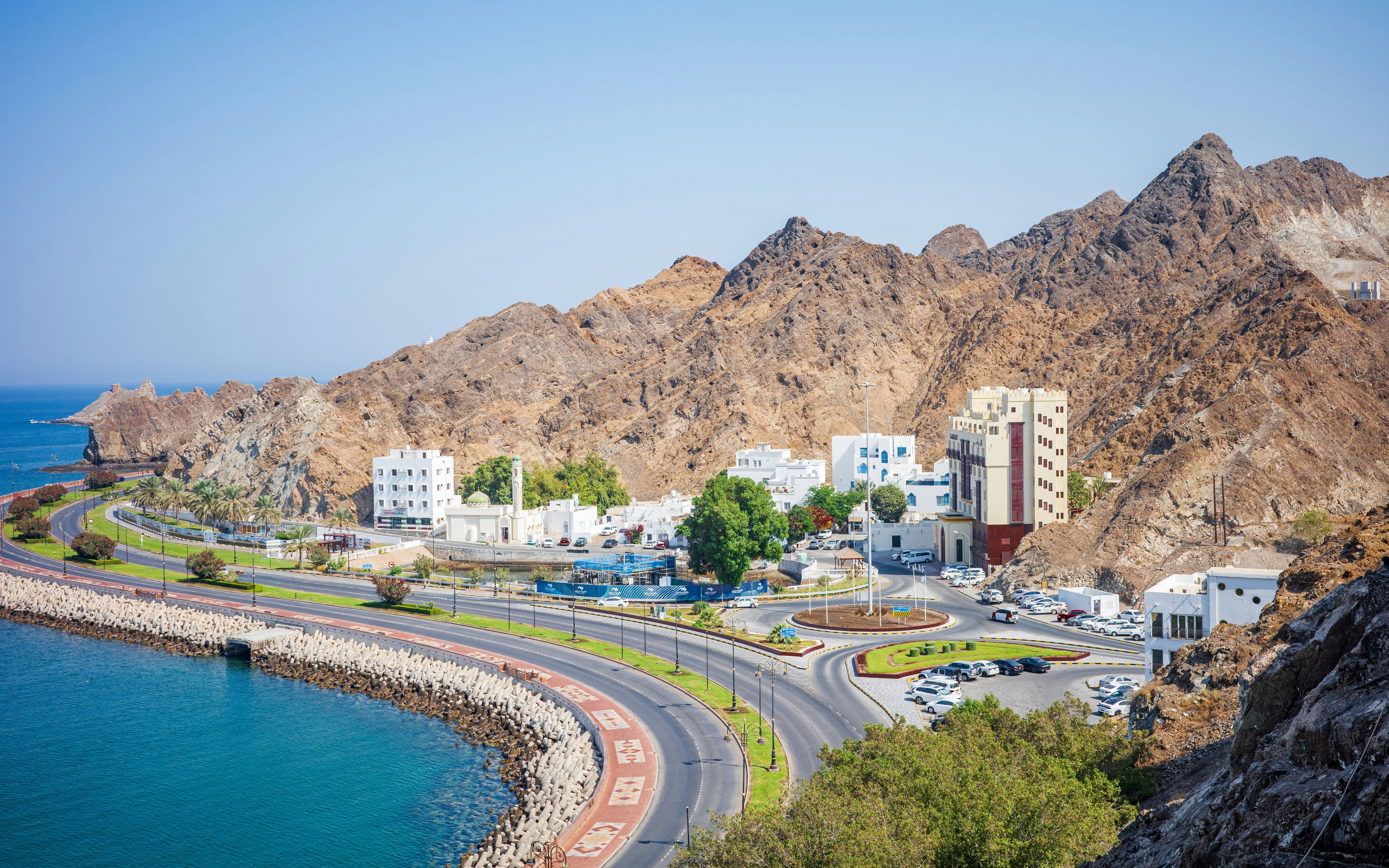 Curving coastal road with traditional white buildings and rugged mountains in Old Muscat, Oman.