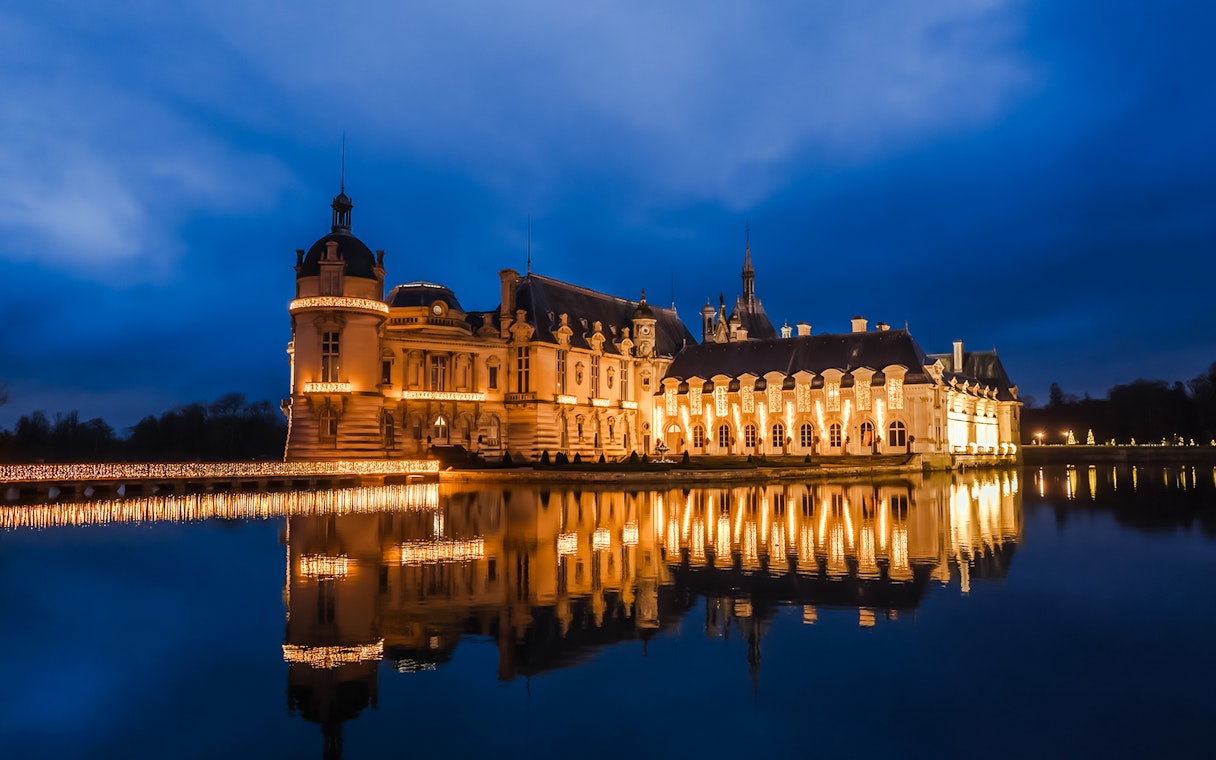 Chateau of Chantilly illuminated at night, reflecting in the water.