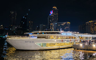 Noah Cruise ship docked at night in Bangkok with city skyline in the background.