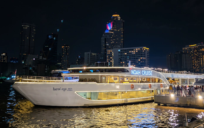 Noah Cruise ship docked at night in Bangkok with city skyline in the background.