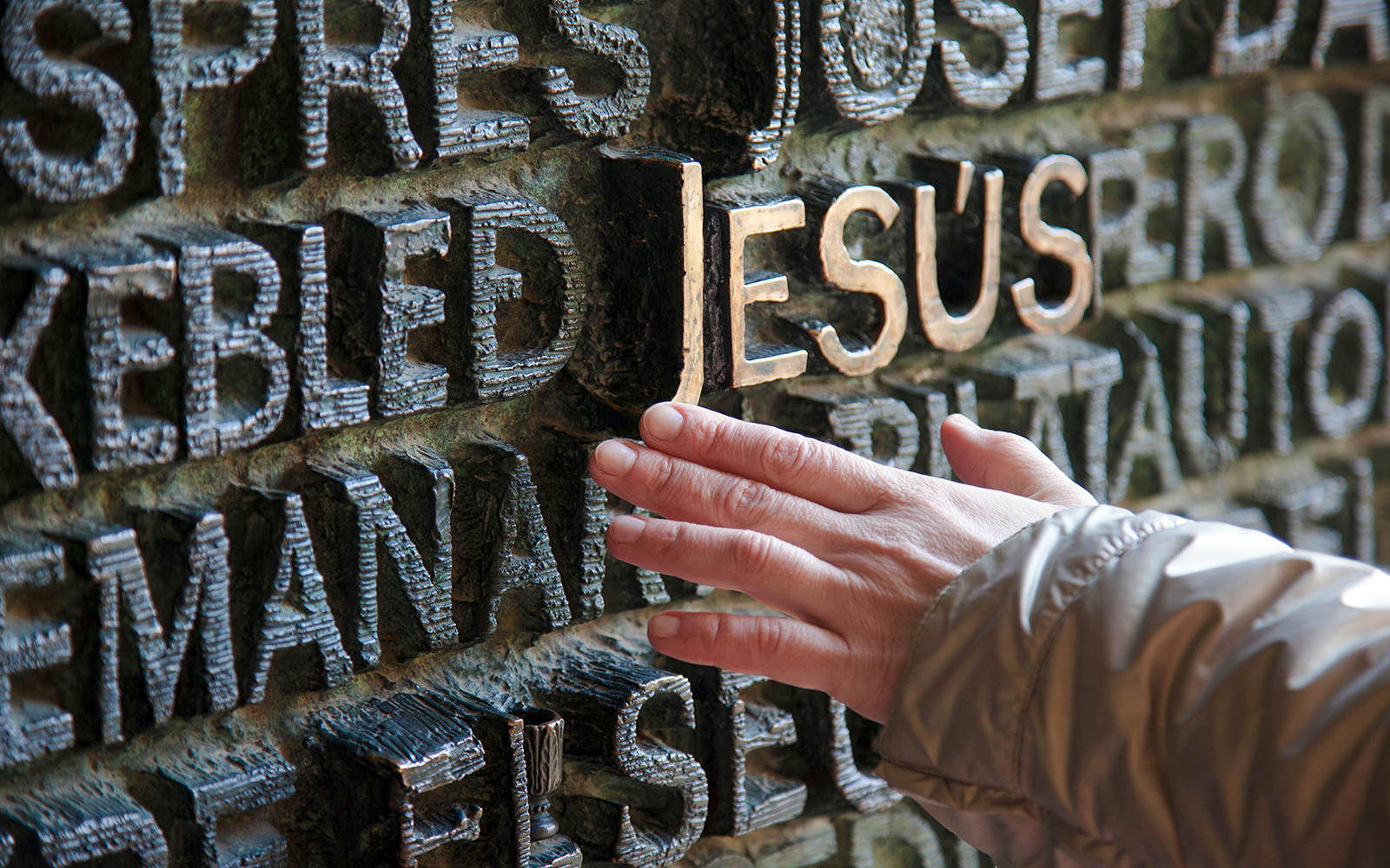 Hand touching ornate bronze doors with raised text at Sagrada Familia, Barcelona.