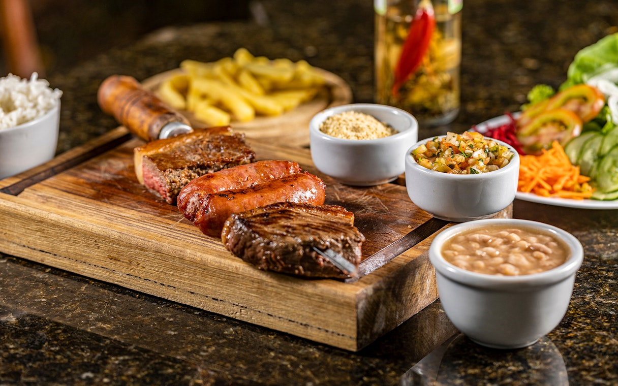 Brazilian barbecue with grilled meats, rice, beans, and sides on a wooden board.