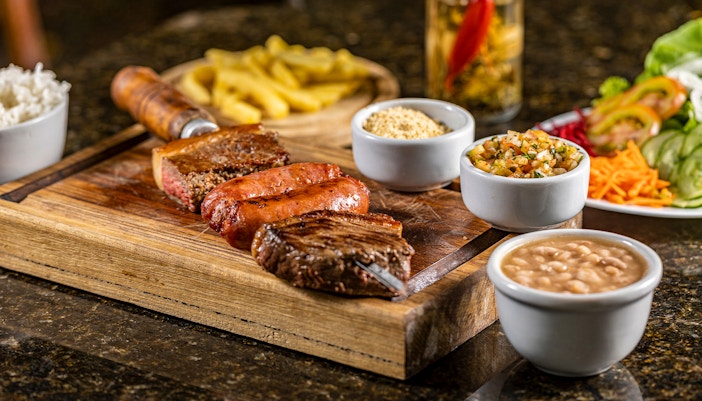 Brazilian barbecue with grilled meats, rice, beans, and sides on a wooden board.