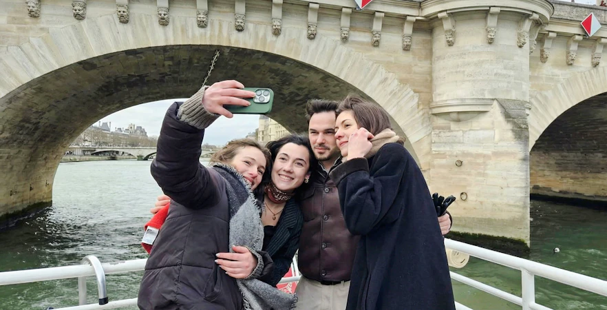 Group taking selfie on Seine River cruise under Paris bridge.
