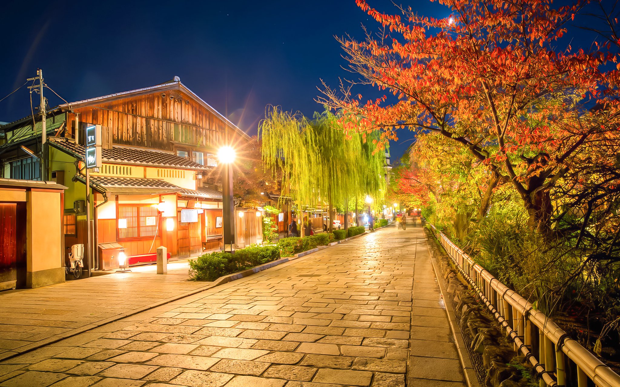 Gion district street in Kyoto, Japan, illuminated at night with traditional wooden buildings and autumn trees.