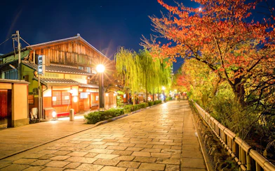 Gion district street in Kyoto, Japan, illuminated at night with traditional wooden buildings and autumn trees.