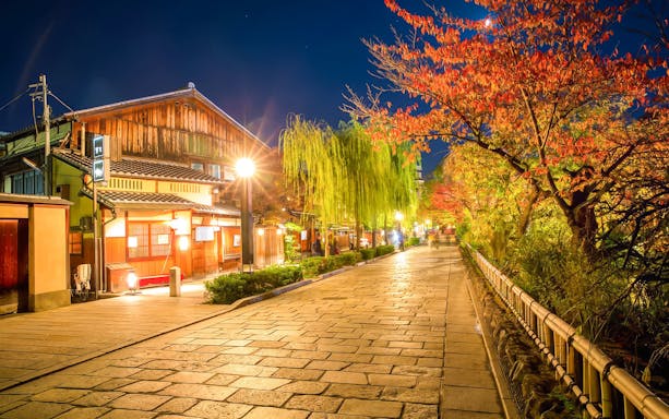 Gion district street in Kyoto, Japan, illuminated at night with traditional wooden buildings and autumn trees.