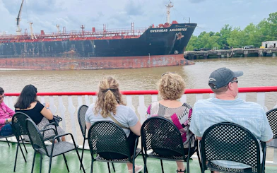 Passengers seated on a riverboat deck watching a large ship pass by in New Orleans.