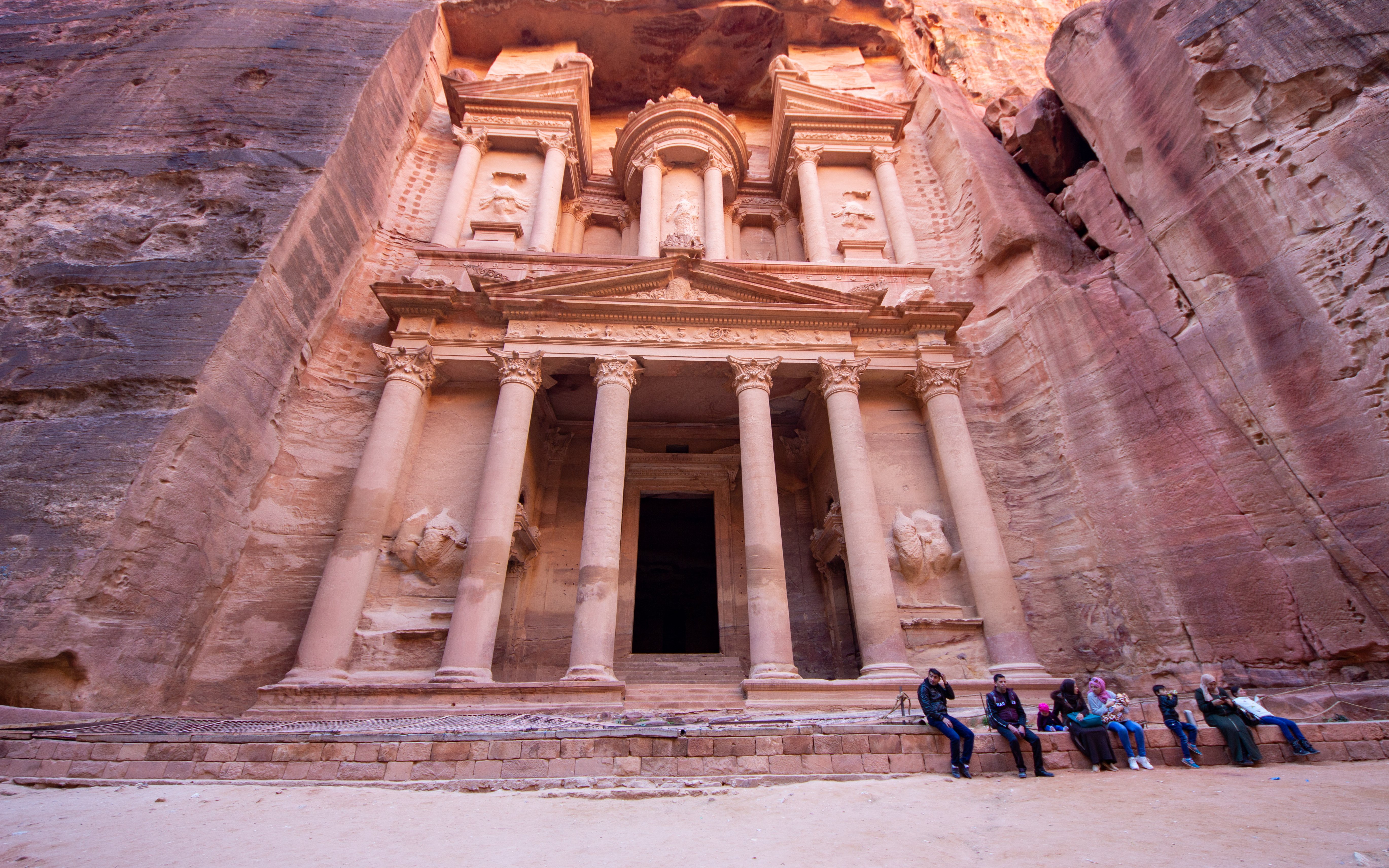 Petra's Al-Khazneh facade with visitors seated, Nabataean archaeological site, Jordan.