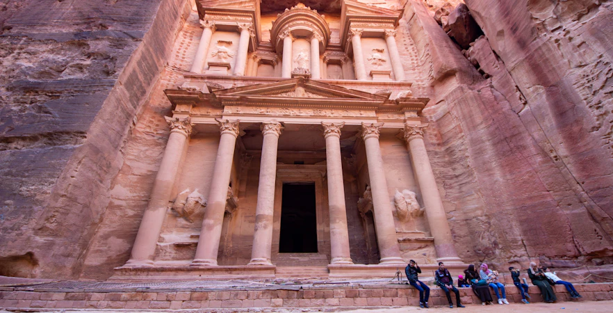 Petra's Al-Khazneh facade with visitors seated, Nabataean archaeological site, Jordan.